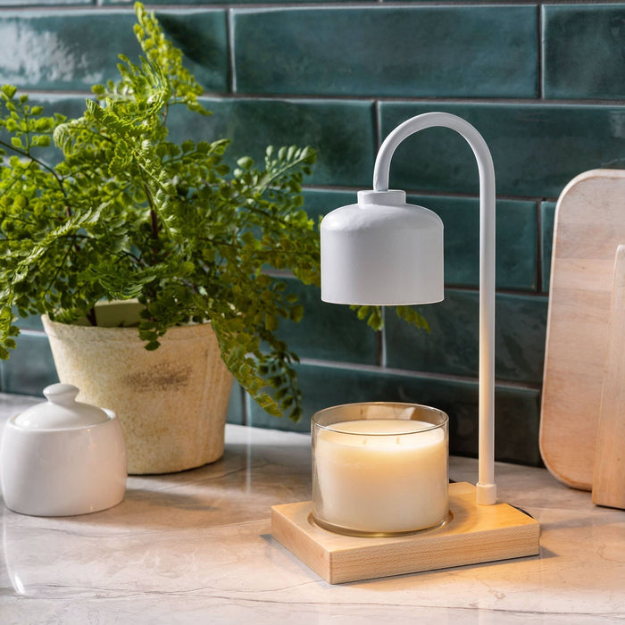 The Polka Dot Press white arched candle warmer lamp with a wooden base and a jar candle on a marble countertop.
